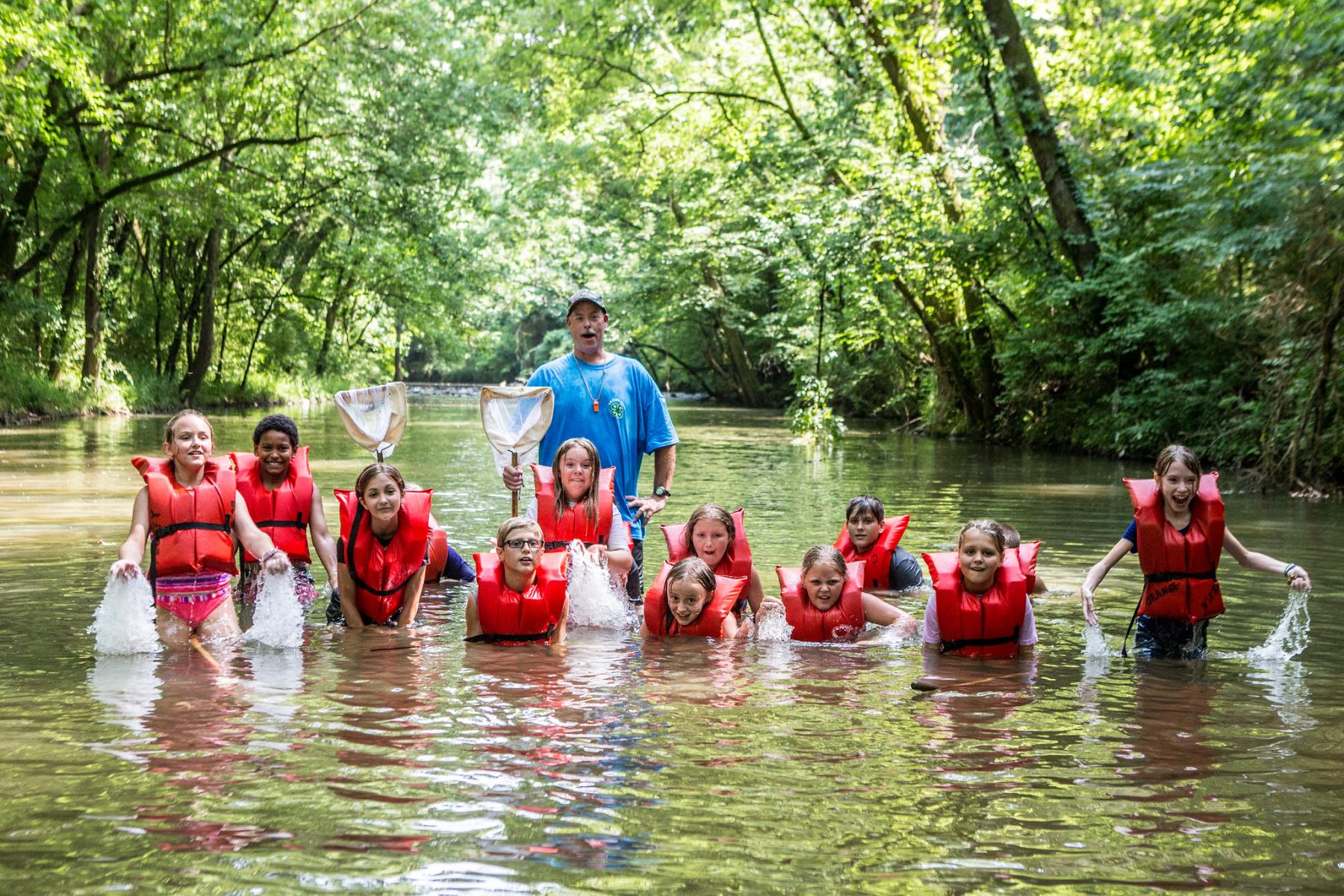adult stands behind children wearing red life jackets playing in river surrounded by green foliage