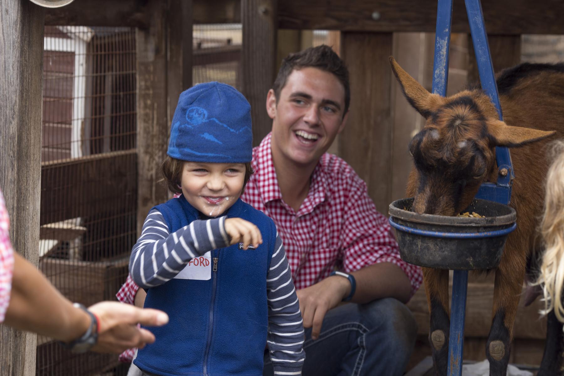 teen boy laughs as a child wipes milk from his lips and walks away from dairy goat.