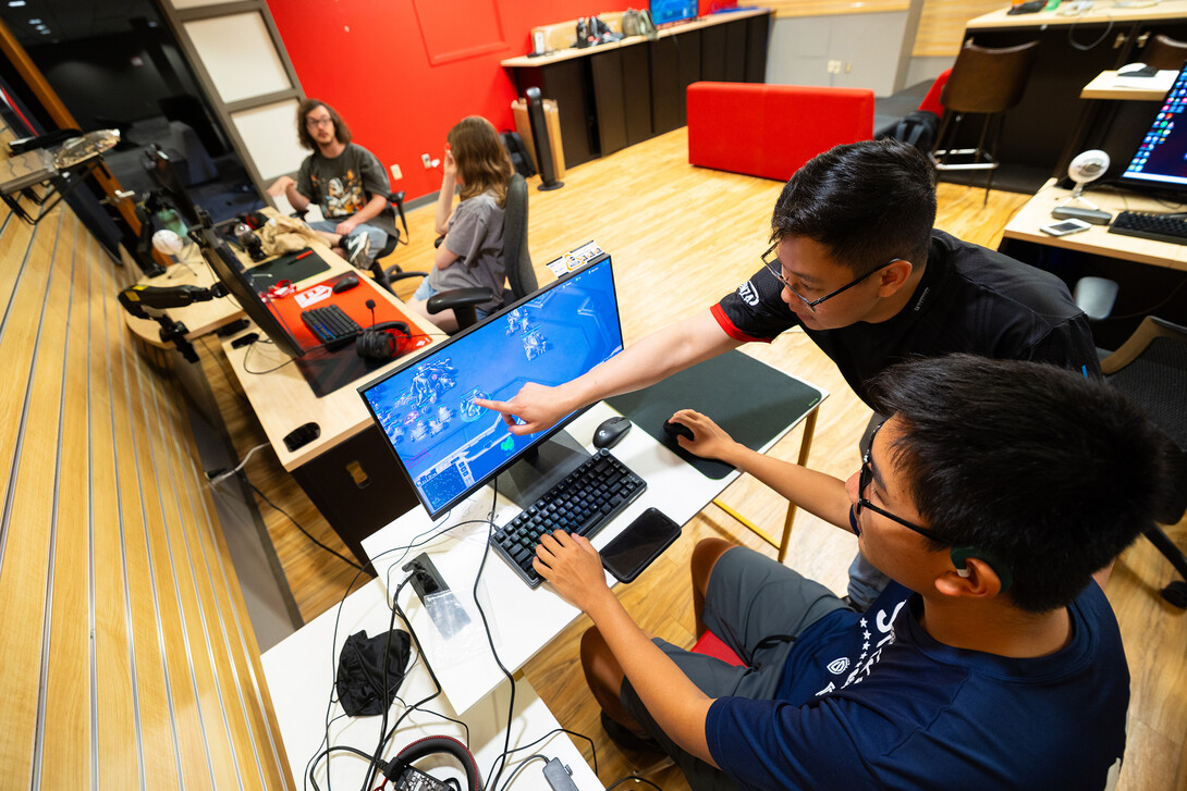 Ryan Tan leans over to point out a spot on a student's monitor as the student sits at the desk playing a game. 
