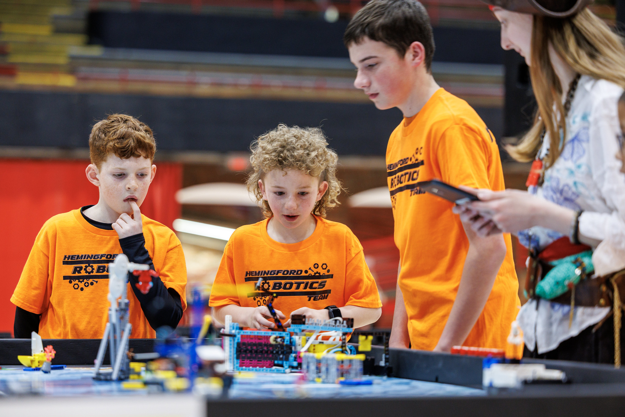 Three youth team members work on a lego robotics project overseen by an attendant in a pirate hat. 