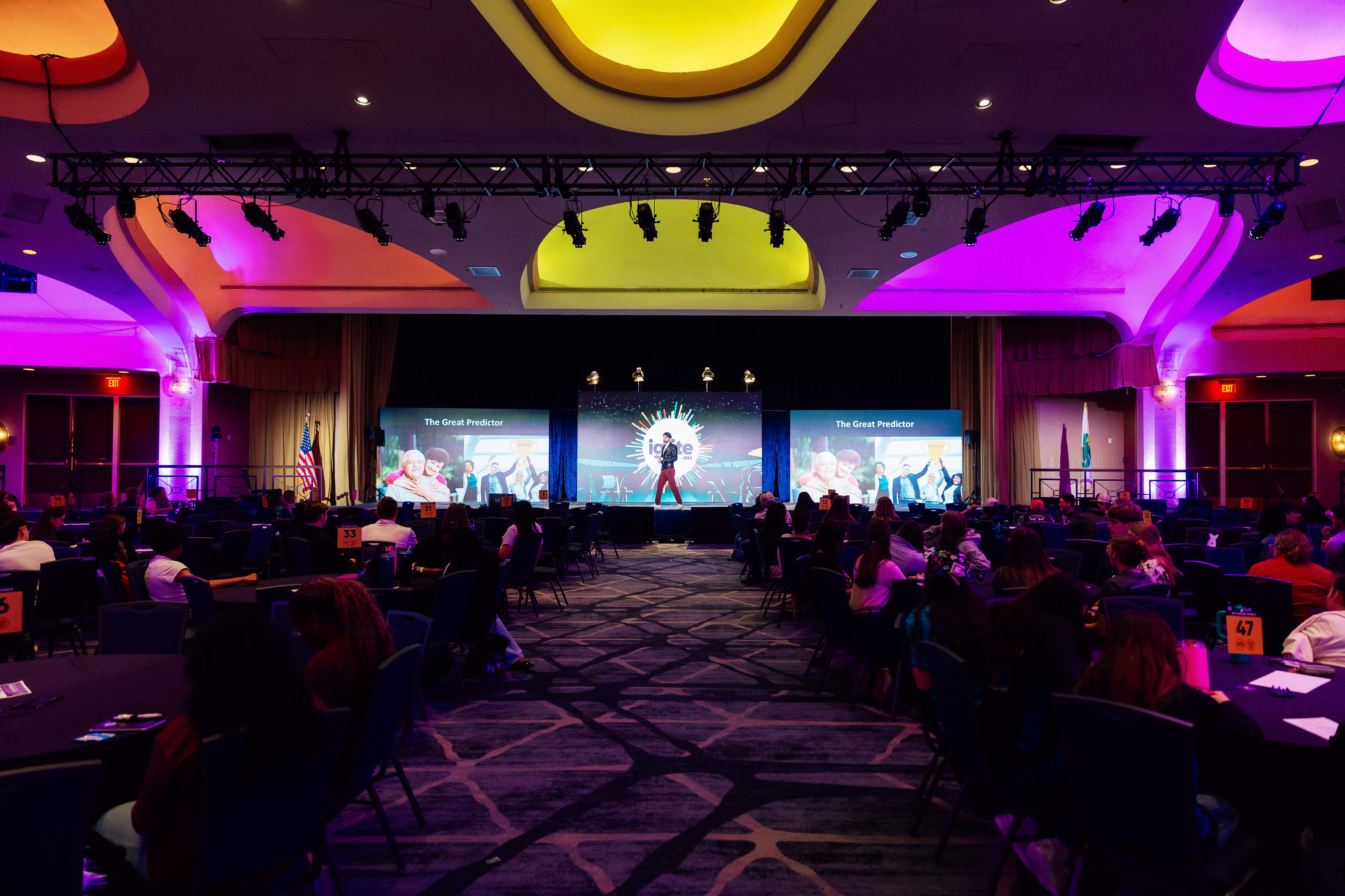 Colorful event hall with a speaker on stage, audience seated at round tables, and vibrant lighting.