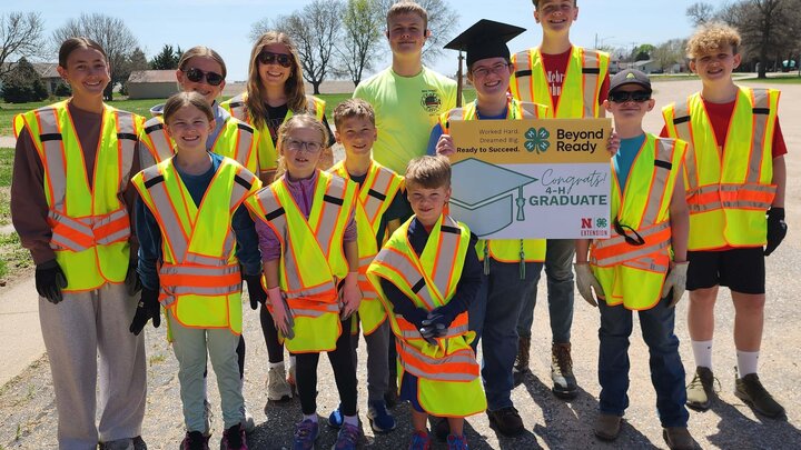 Group of people wearing bright safety vests outdoors, holding a graduation-themed sign.