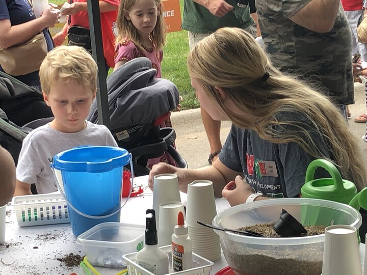 A Thrive Lab intern at East Campus Discovery Days helps a child plant grass seeds and make a project to take home and grow. 